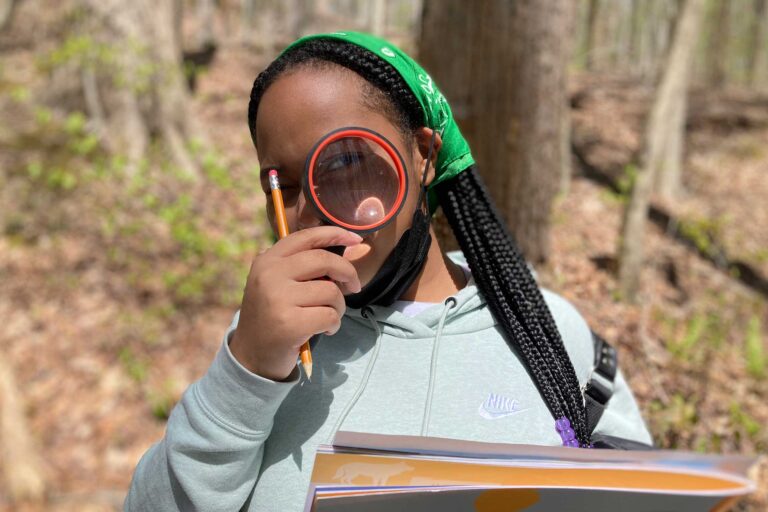 a young student in the woods holding a book and looking through a magnifying glass