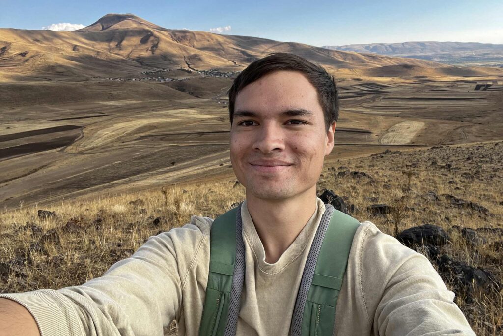 a young man standing on a hillside photographing himself with mountains in the background