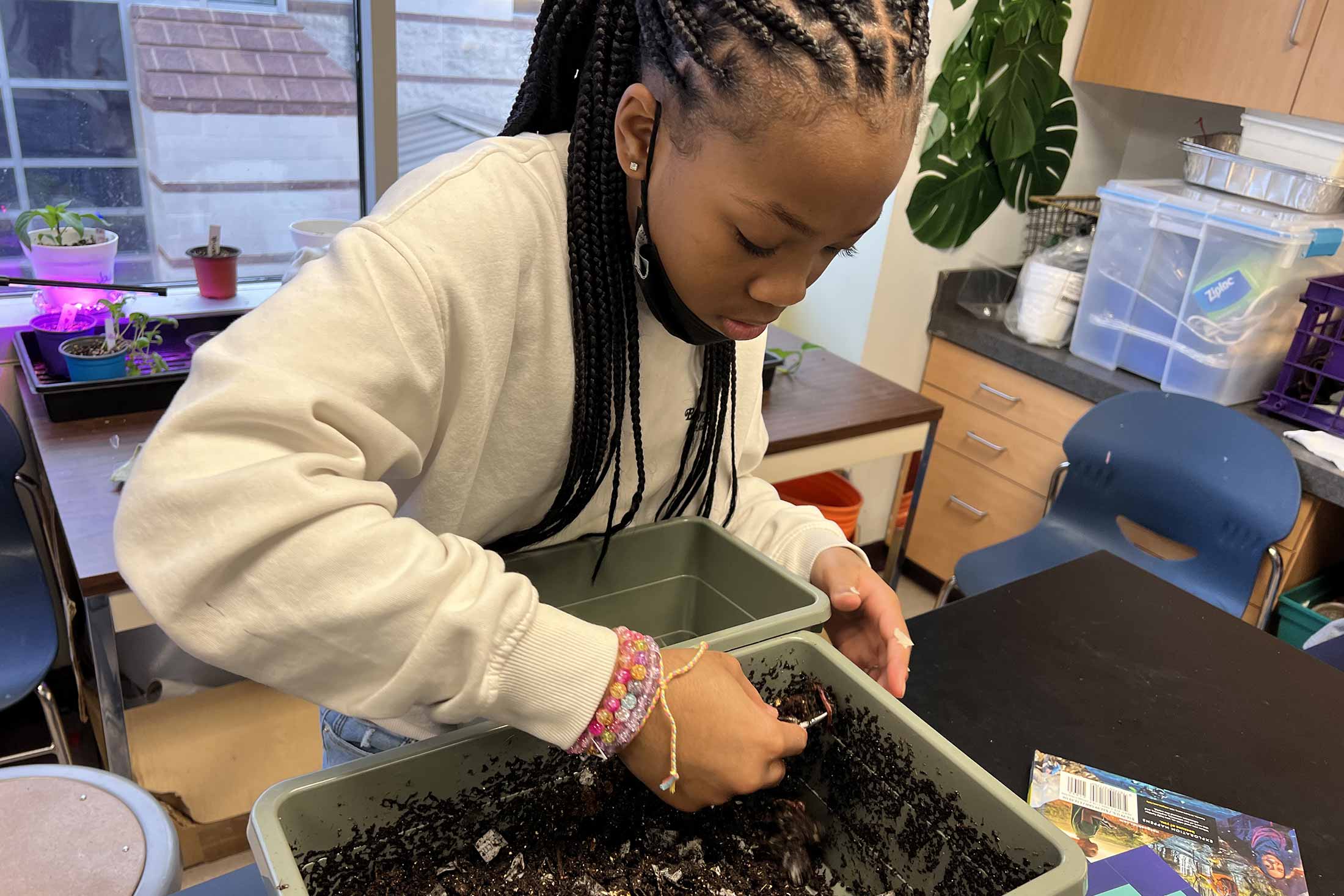 a young Black female student composting on a table in a school classroom with some plants in the background