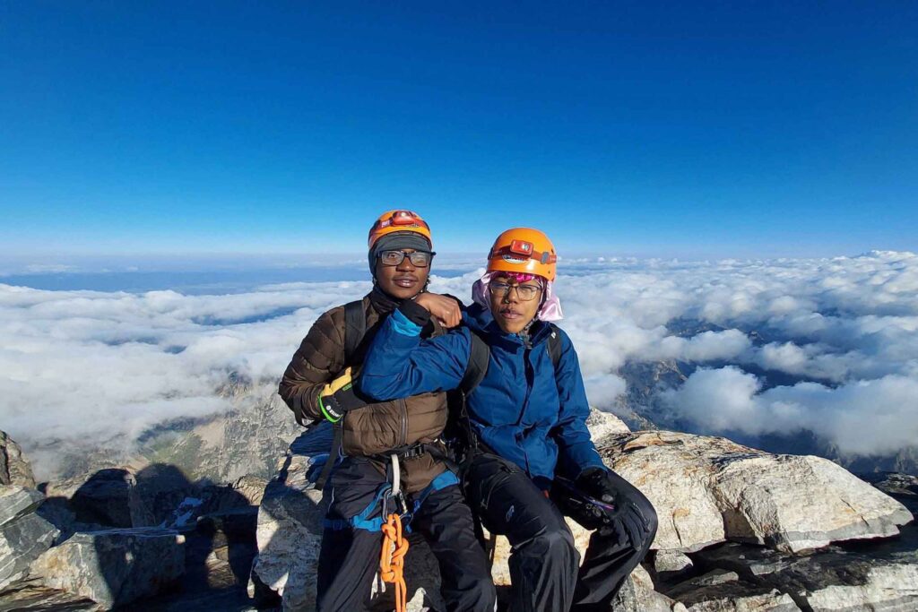 two young Black teenagers wearing mountain climbing gear sitting on a boulder above the clouds