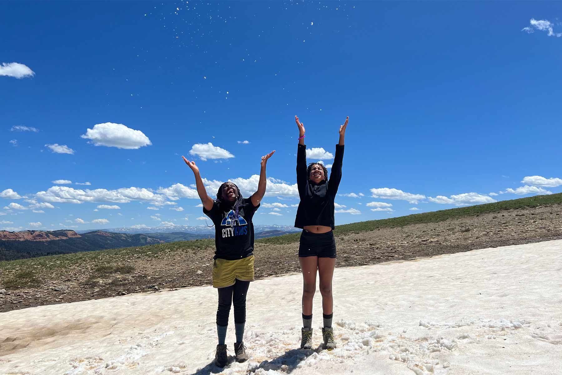 two young Black female teenagers on a hillside smiling while throwing snow up in the air