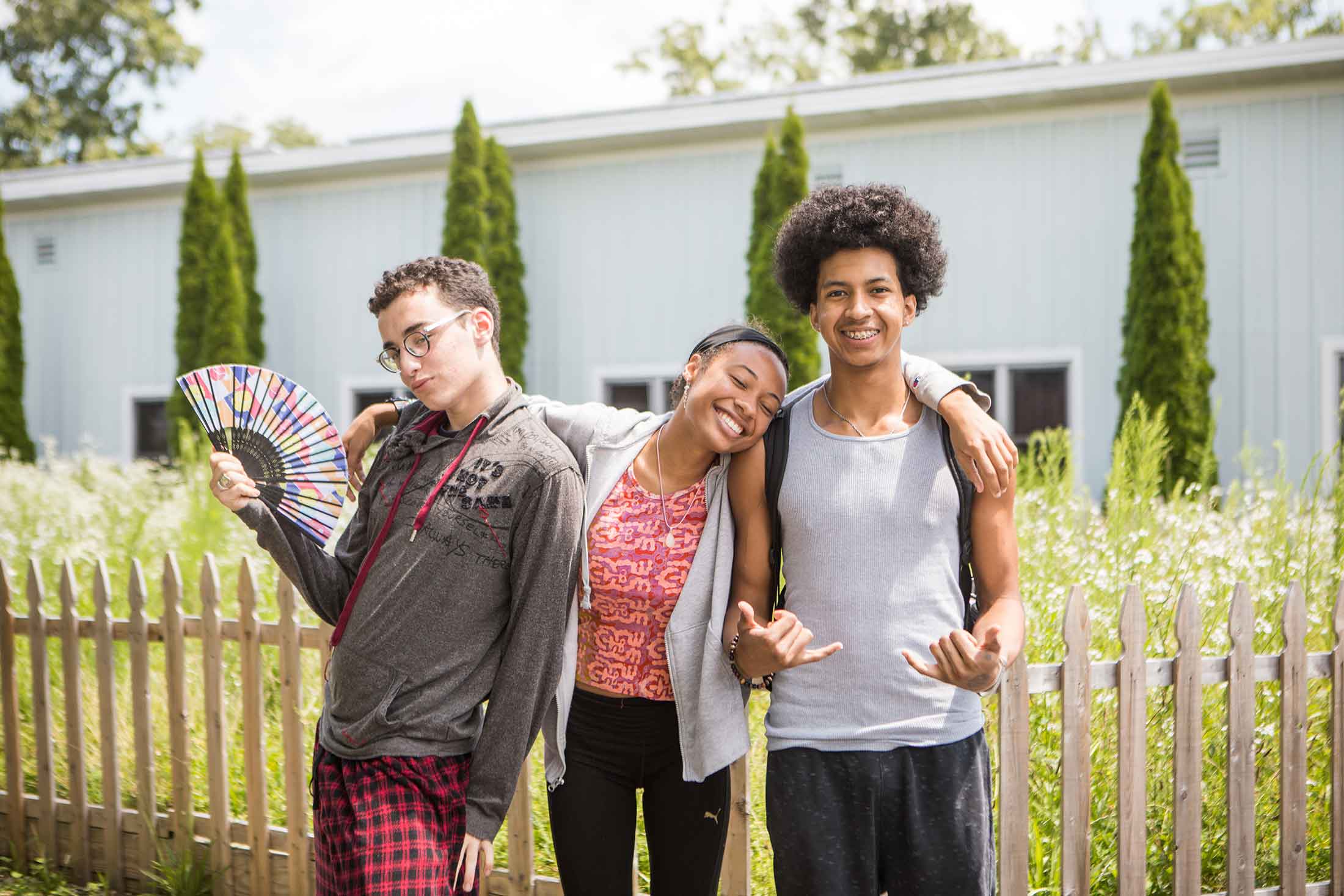 a group of three young people, one holding a colorful fan, smiling while posing for a picture in front of a wood fence with trees and a building in the background