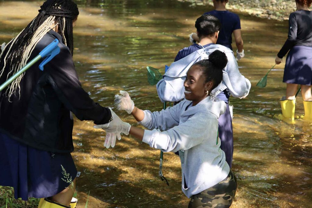 a group of young students wearing yellow waterproof boots and holding small green nets while walking in a creek