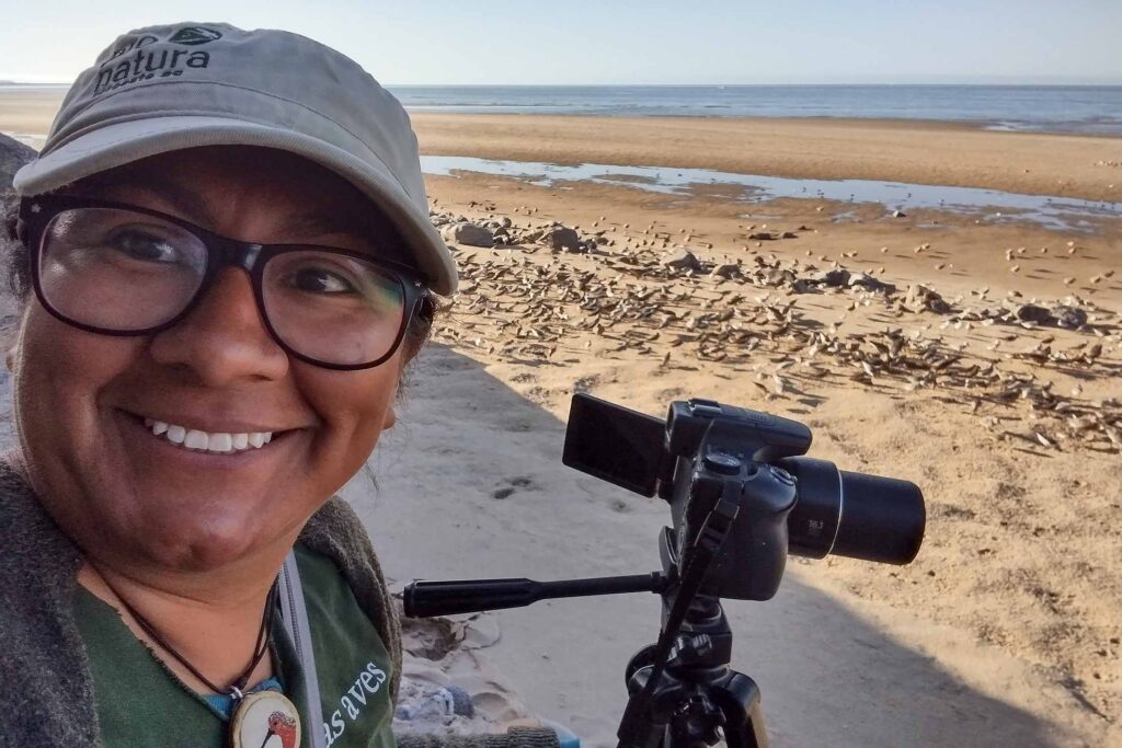 woman wearing a hat and glasses taking a selfie with a beach and water in the background