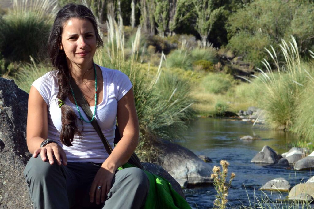 a woman wearing white shirt and turquoise beaded necklace sitting on a large rock at the edge of a stream