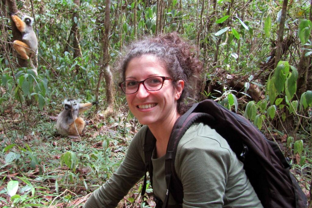 a woman wearing glasses, a green shirt, and dark backpack kneeling in a jungle with two brown, gray, and black primates in the background