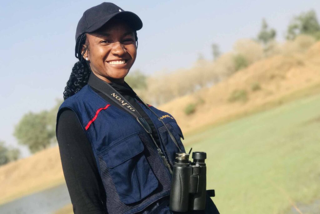 a Black woman wearing a baseball cap and blue jacket with binoculars around her neck smiling with green and tan foliage in the background