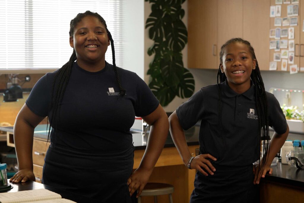 two young Black school girls wearing blue shirts standing in a lab speaking to the camera