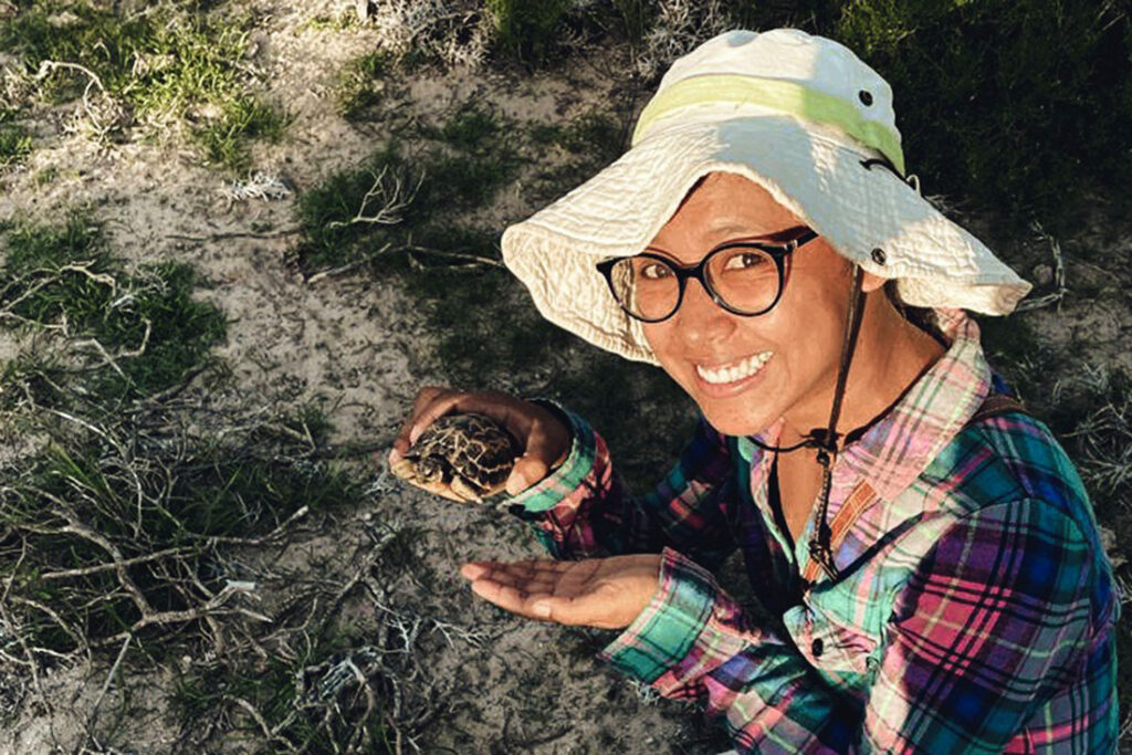 a woman wearing glasses, a hat, and pastel colored shirt smiling while holding a small turtle