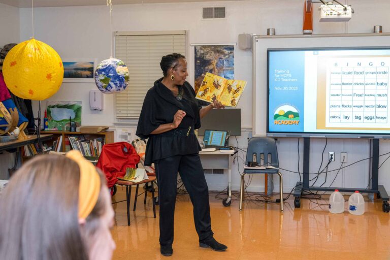 a Black woman holding a book and reading in front of a classroom