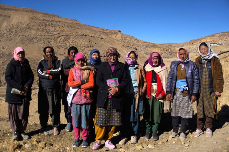 a group of ten women wearing cold weather clothes and head scarves standing on rocky ground with mountains in the background