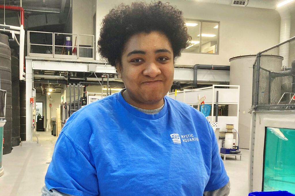 a young Black woman wearing a blue shirt with a Mystic Aquarium logo smiling while standing in a lab