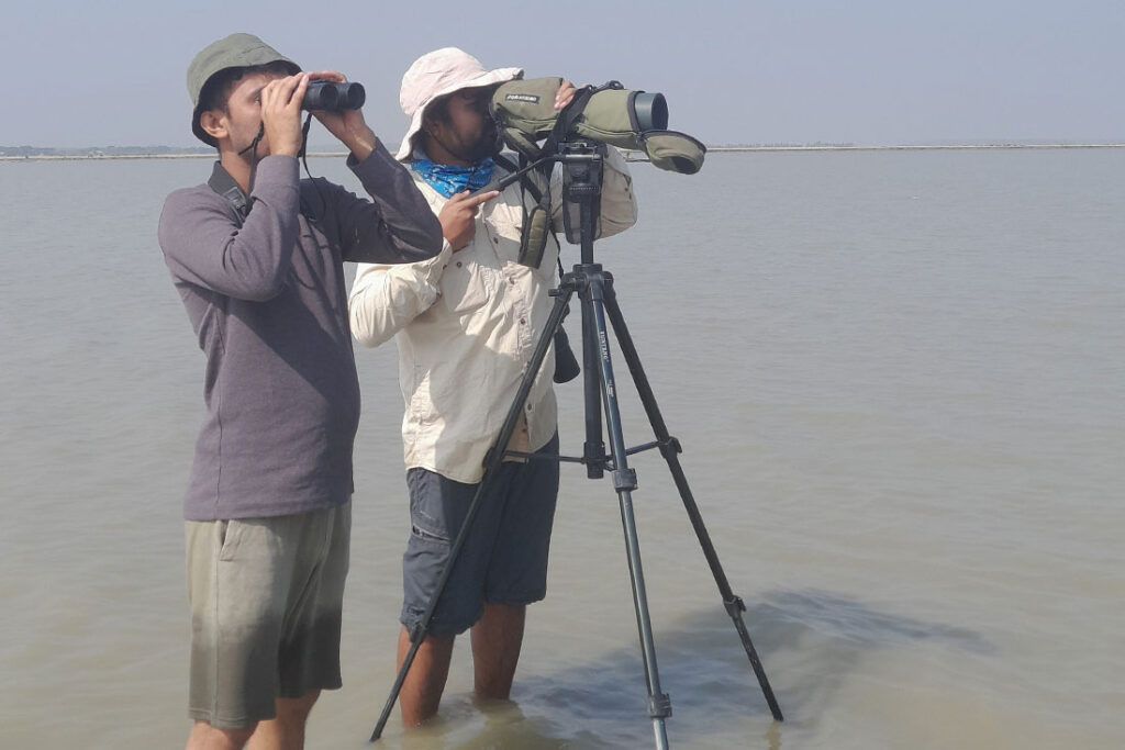 two young men standing in shallow water; one holding binoculars and one looking through a lens situation on a tripod