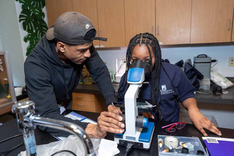 a young Black man wearing a blue hooded sweatshirt and baseball cap assisting a young Black female student as she looks through a microscope in a classroom lab