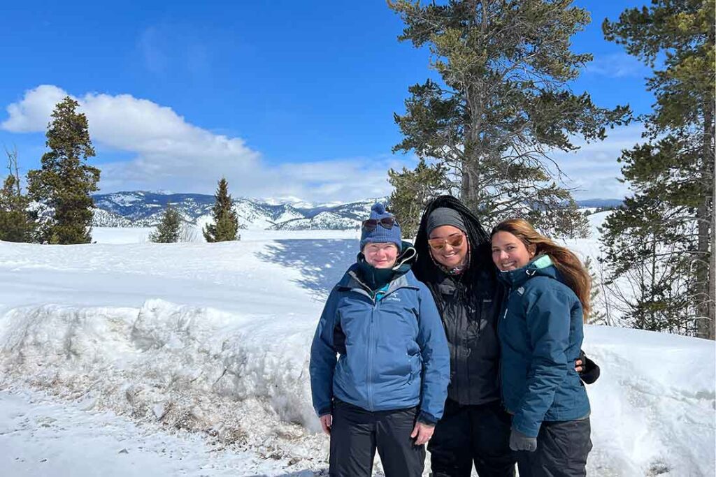 three women wearing cold weather gear in the snow with snow covered mountains in the background