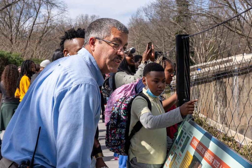 a Black man wearing a blue collared shirt and classes looks through a fenced enclosure at the zoo alongside young Black students