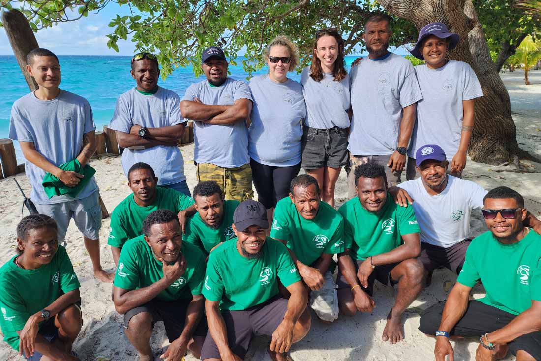 a group of men and women wearing t-shirts and posing on the sane under a tree with bright blue water in the background
