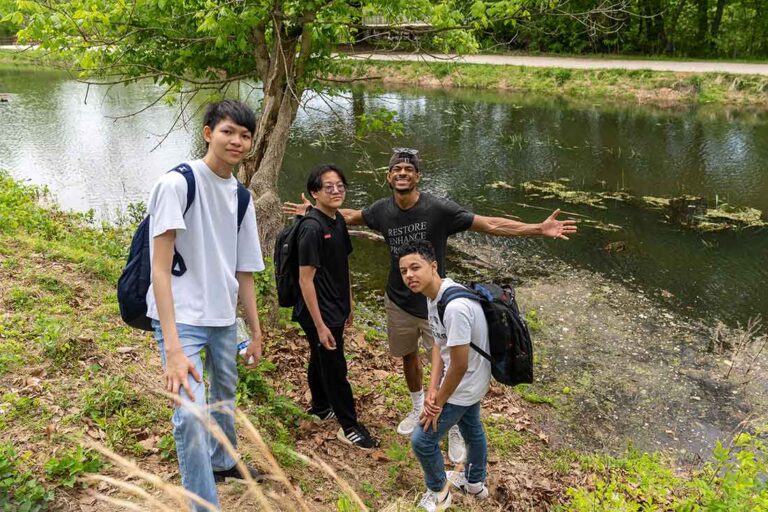 a young black man with outstretched arms poses for a photo with three young teens at the bank of a canal