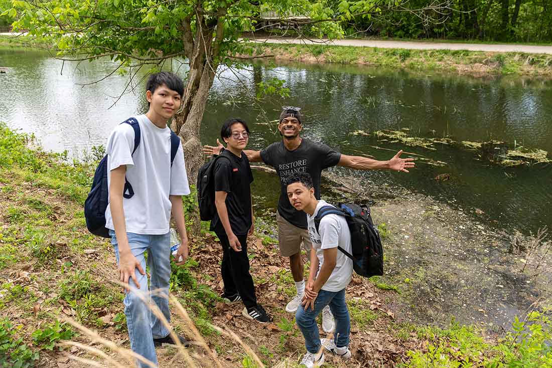 a young black man with outstretched arms poses for a photo with three young teens at the bank of a canal