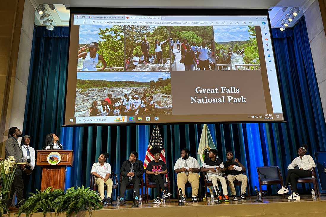 a group of people standing at a podium looking at a presentation on a projection screen with other people sitting up on a stage