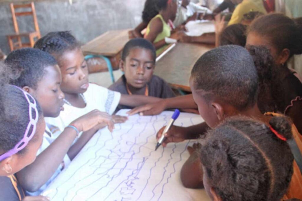 a group of small children sitting around a desk look at a large piece of paper as one student draws on it