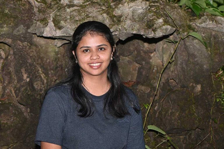 a woman with dark hair wearing a blue shirt stands in front of a large rock