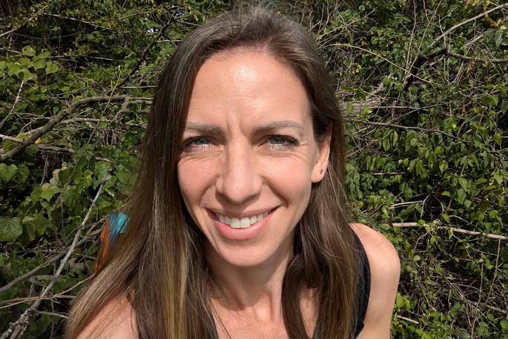 a woman with light brown hair takes a selfie while standing in front of some green shrubs