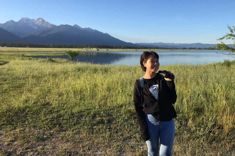 a woman wearing a backpack and holding binoculars standing near a body of water with mountains in the background