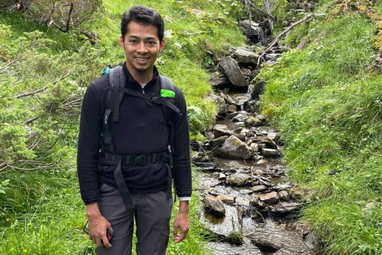 a man wearing a jacket and backpack standing in grass next to a shallow stream