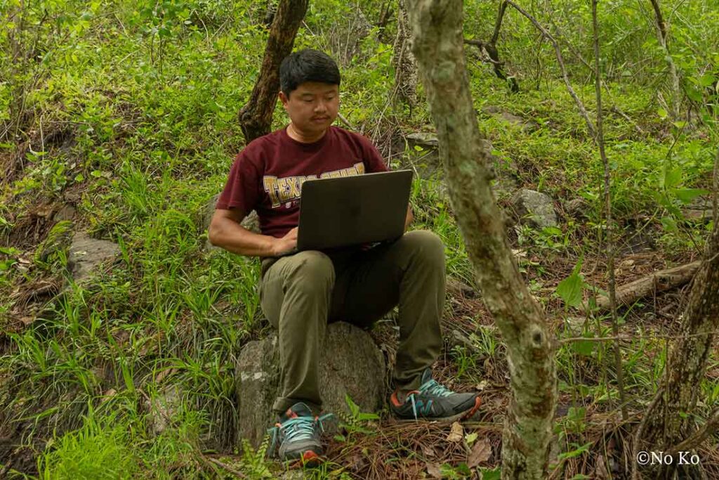 a man with dark hair sits on a rock in the woods while working on a laptop computer