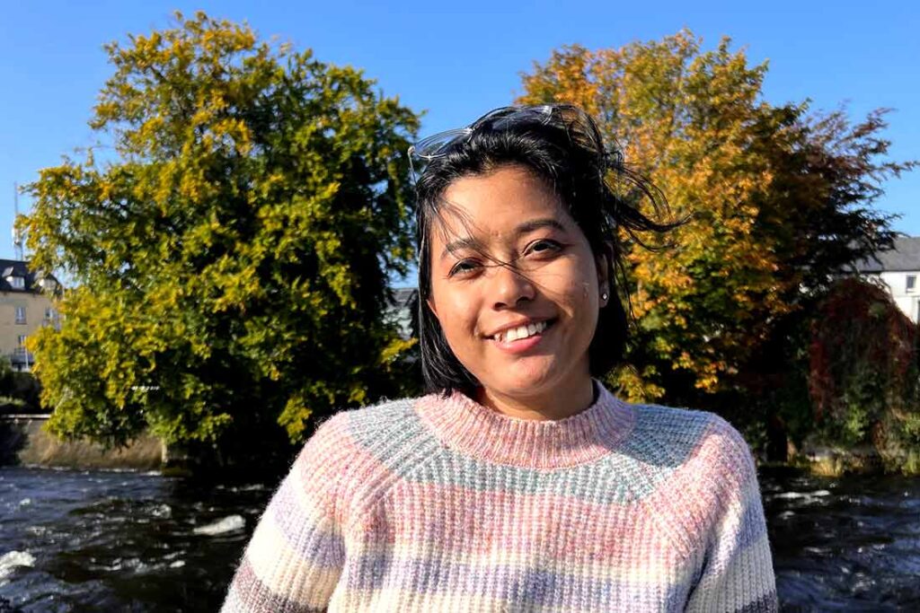 a woman with dark hair wearing a colorful sweater poses with trees and buildings in the background