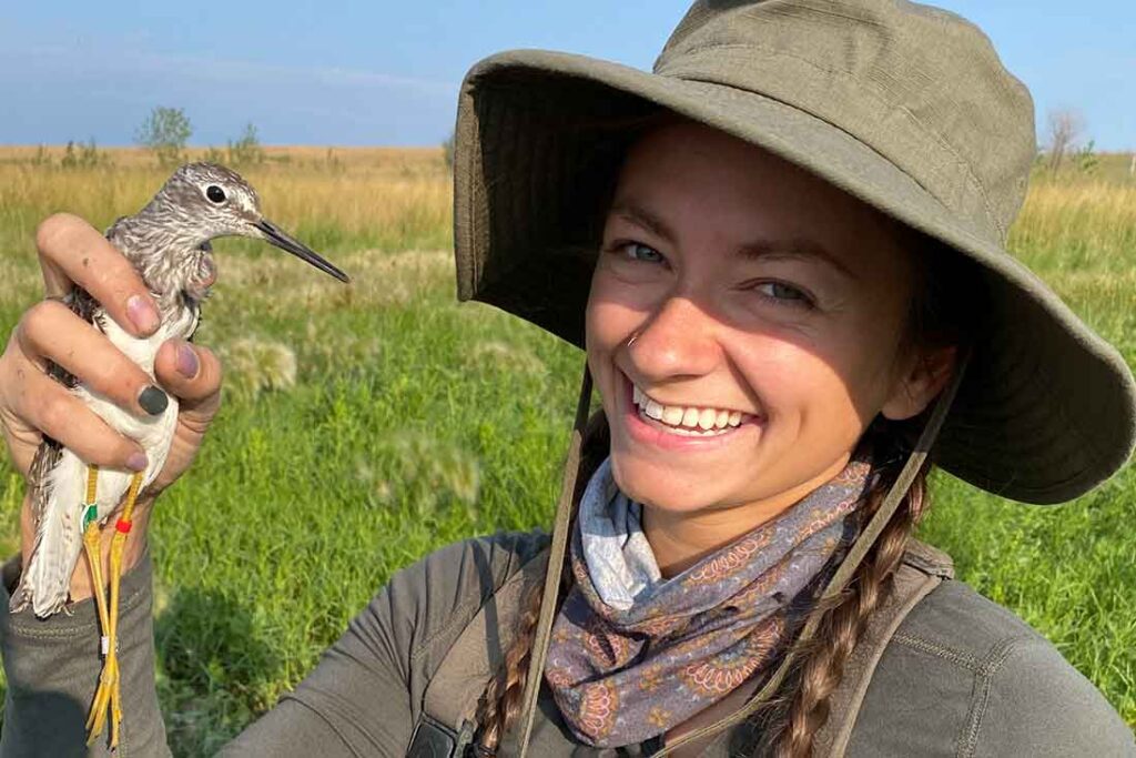 a woman wearing a green hat smiles while holding a shore bird with identification bands on its long yellow legs