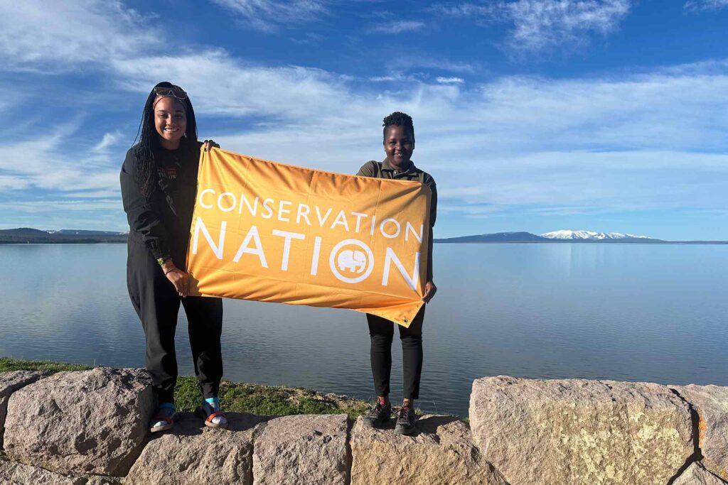 two Black women holding an orange flag that reads conservation nation, while standing on a rock structure with water and mountains in the background