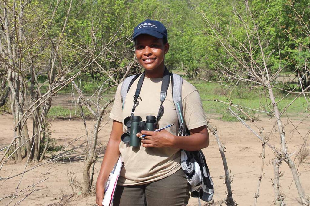 a Black woman wearing a blue baseball cap and a backpack with binoculars around her neck standing on dirt with green trees in the background