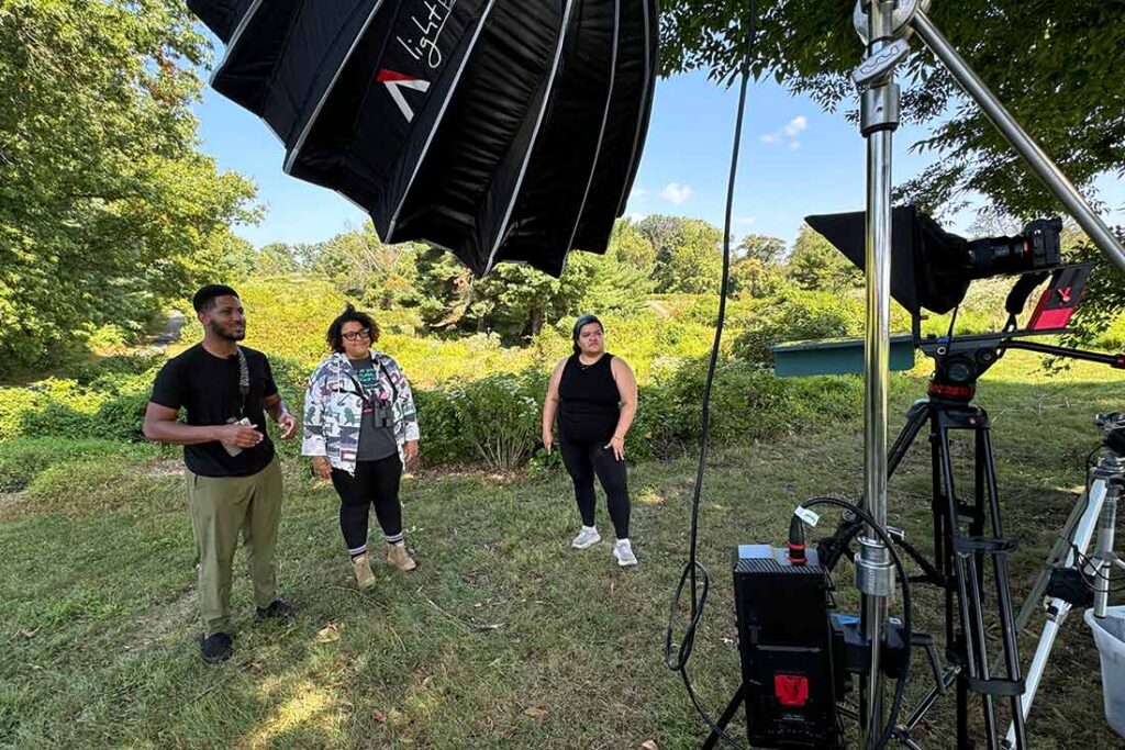 three people standing outside in the grass with trees in the background looking at video production equipment