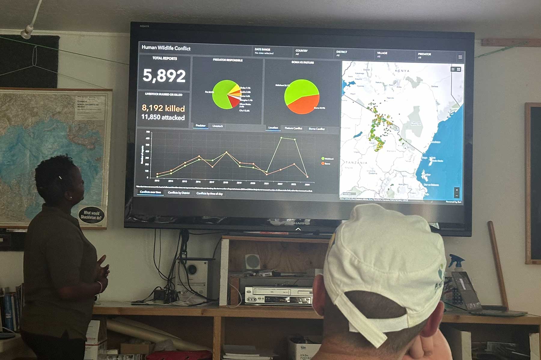 a Black woman in an office presenting data on a large screen while a person in the foreground listens