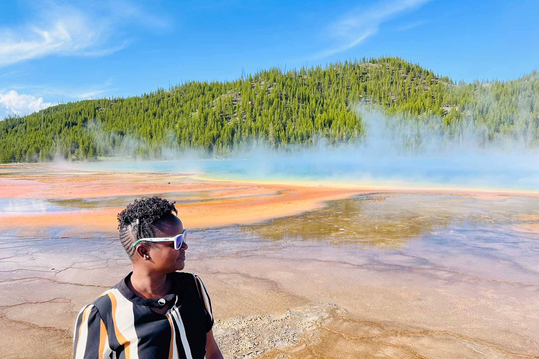 a Black woman standing near a hot spring with trees in the background