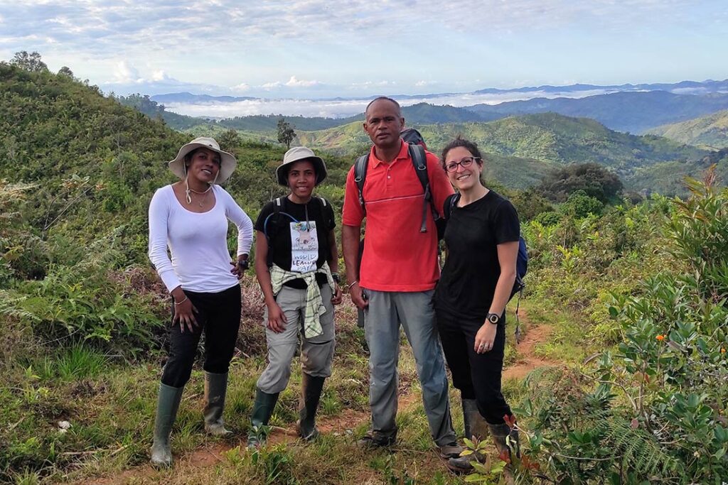 four people standing on grass and dirt at a high elevation with mountains in the background