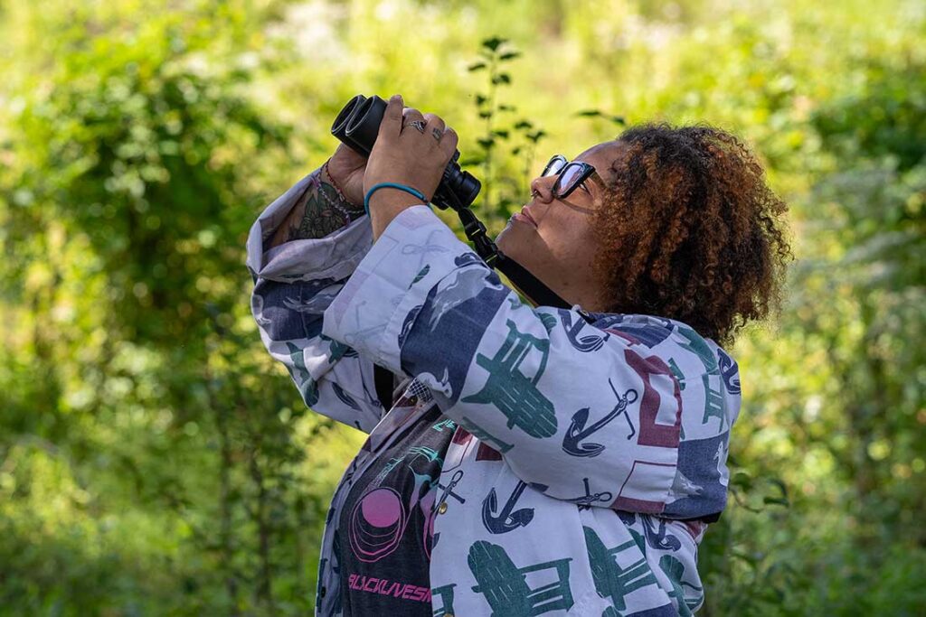 a Black woman with curly hair wearing glasses uses binoculars to look up, with green foliage in the background