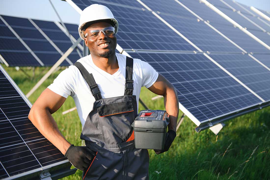 a Black man wearing safety gear while holding a toolbox stands in a grass field with solar panels in the background