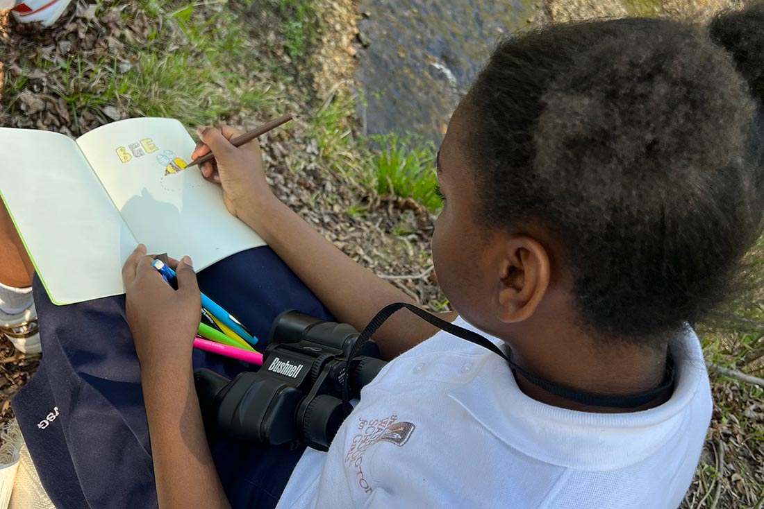 a Black girl wearing a school uniform and binoculars around her neck drawing with colored pencils while sitting outside