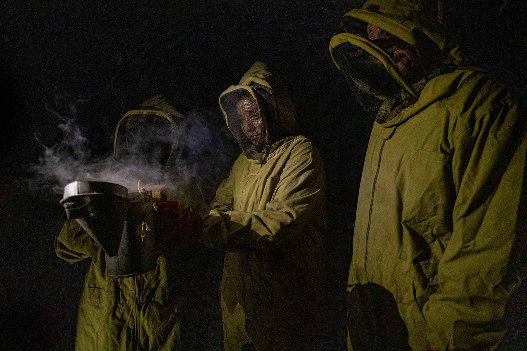 three people dressed in bee protective gear using a specialized smoke machine in the dark