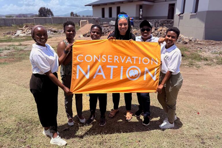 six Black people standing in a grass lot in front of a building while holding a large orange banner that reads conservation nation