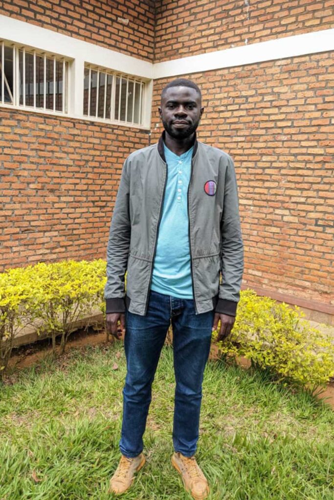 a Black man wearing a light blue shirt, gray jacket, and jeans poses while standing on grass in front of a red brick building