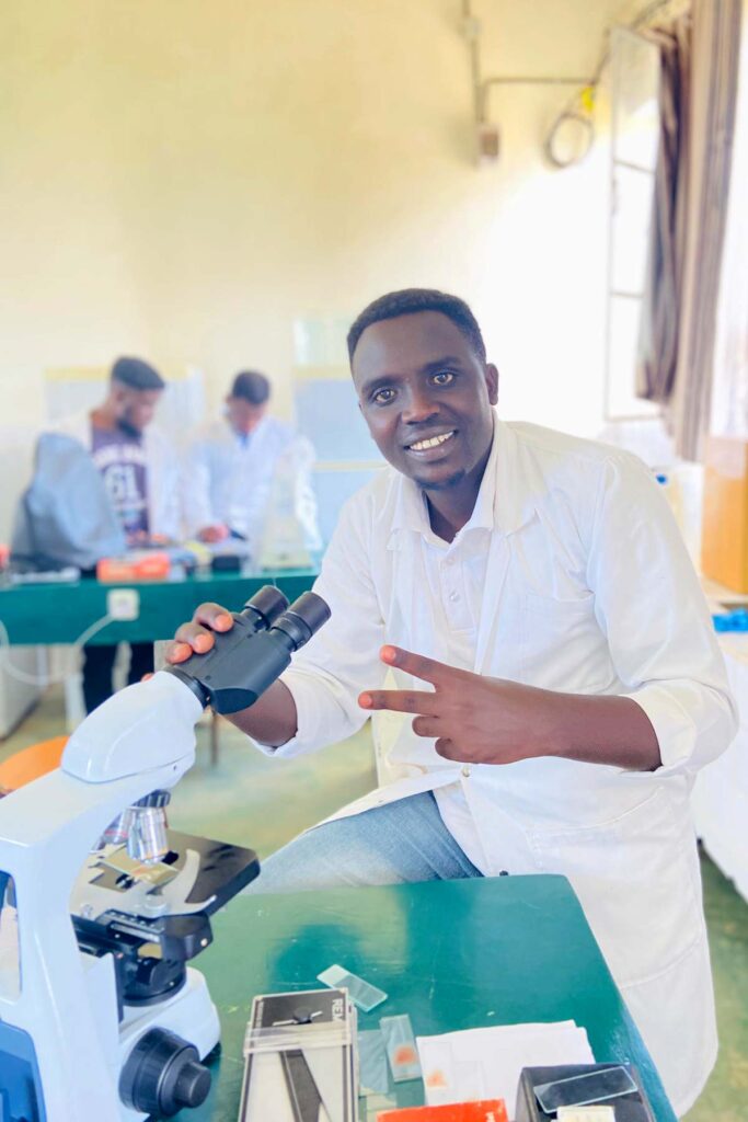 a Black man wearing a white lab coat poses while using a microscope in a lab