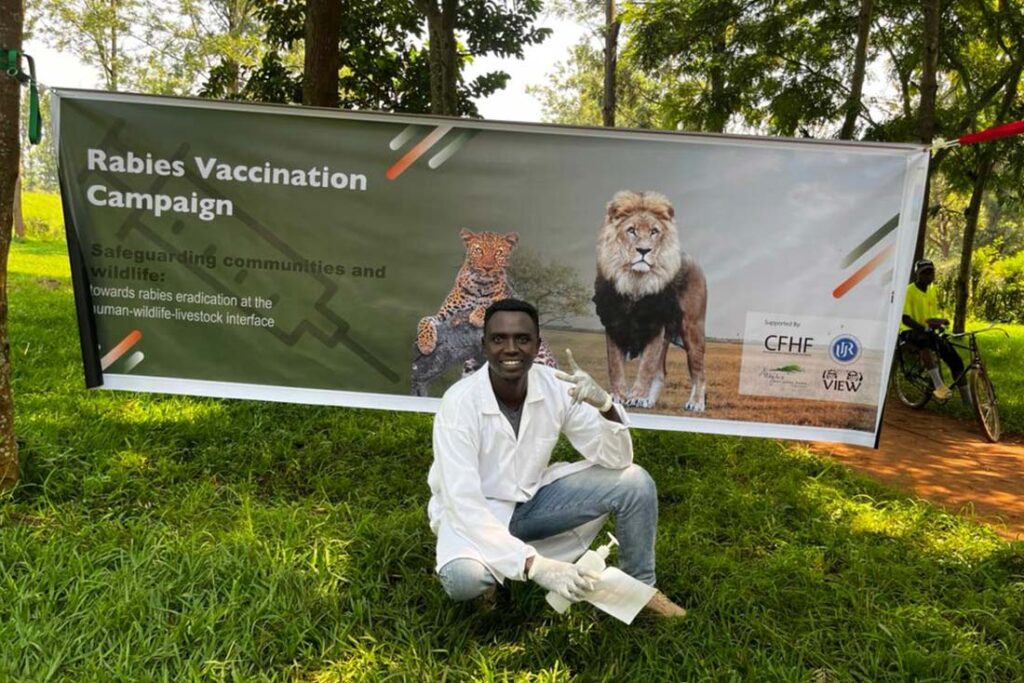 a Black man wearing a white medical coat and gloves poses next to a sign about a rabies vaccination campaign