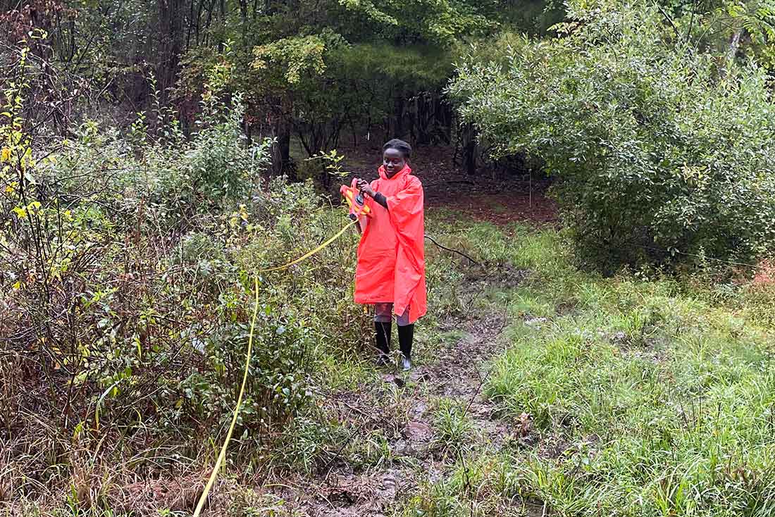 a Black woman wearing a red rain poncho using a yellow tape measure while standing in a wooded area