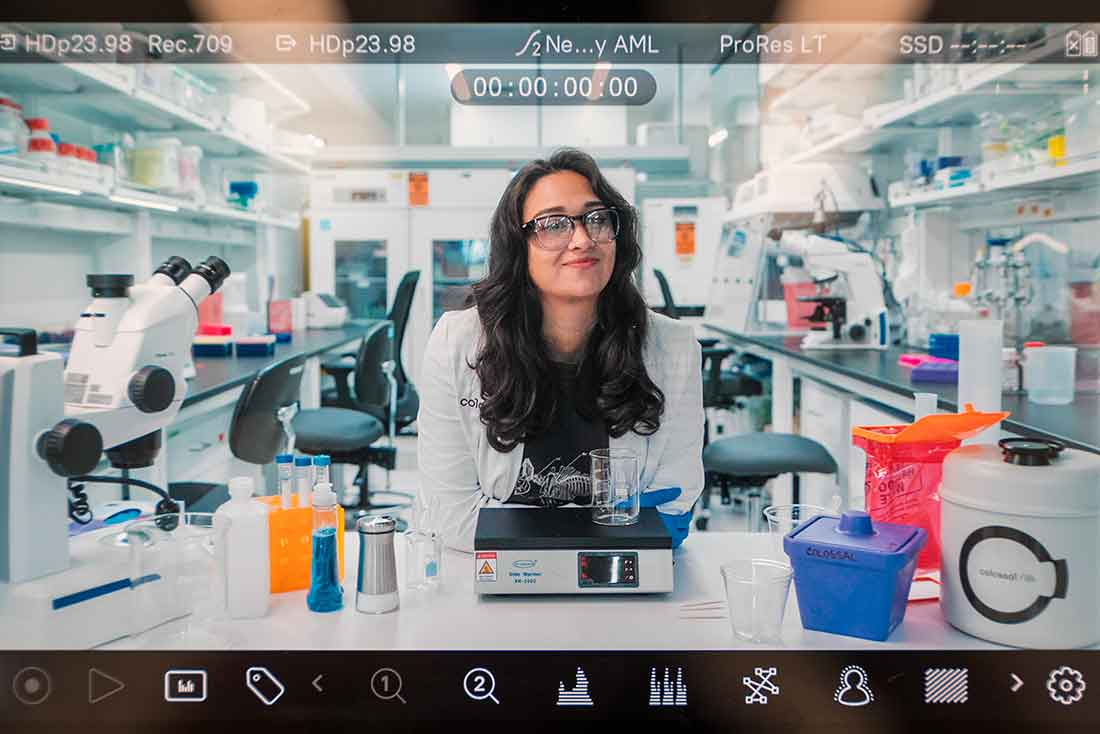 A scientist in a lab coat smiles confidently in a bright, organized laboratory. Surrounding her are microscopes, colorful liquids, and equipment, conveying a sense of innovation and focus.