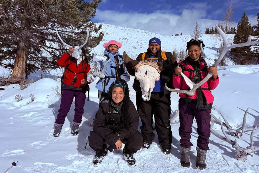 Five people in winter gear smiling in a snowy landscape, holding antlers and a skull. Bright blue sky and snow-covered hills in the background.