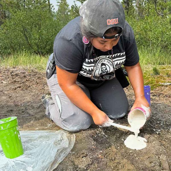 a woman wearing a baseball cap in the field pouring plaster into an animal track on the dirt